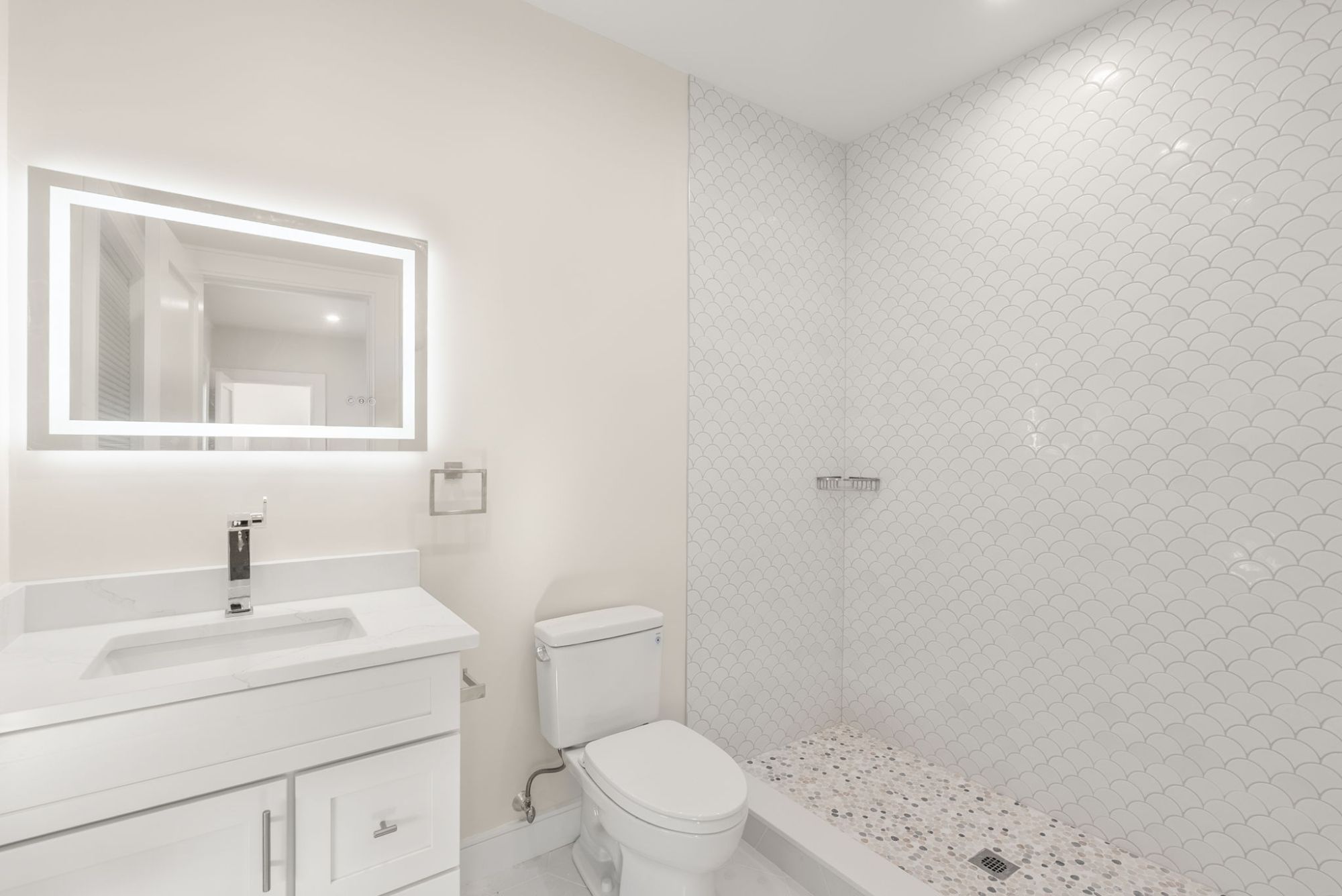 Bathroom with a white scallop-tile shower, pebble floor and lit mirror